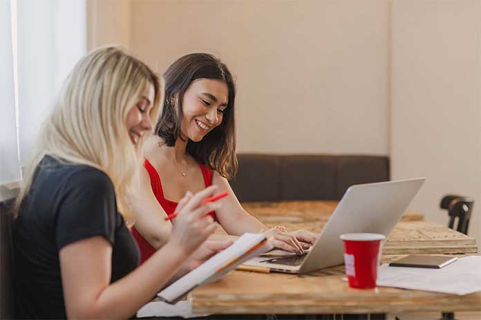 Two people sitting at a table smiling, one is typing on a laptop and the other is holding a pen and notebook
