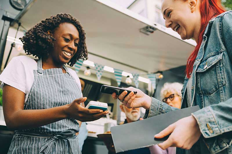 A person standing in front of a food truck wearing an apon taking a card payment from a customer smiling A person standing in front of a food truck wearing an apon taking a card payment from a customer smiling