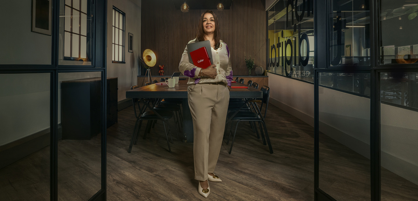 Surinder standing in her office with a notebook and laptop in one arm. Surinder standing in her office with a notebook and laptop in one arm.