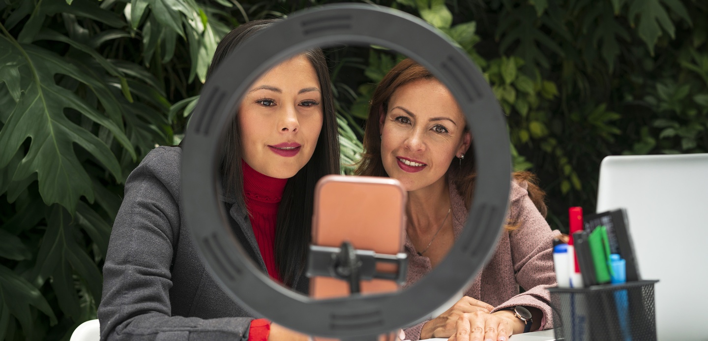Two women sat at a desk, framed by a ring light, looking at a smartphone recording content.