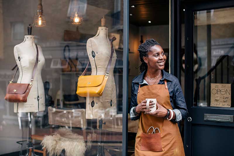 Person wearing an apron standing in front of a shop smiling and holding a mug Person wearing an apron standing in front of a shop smiling and holding a mug