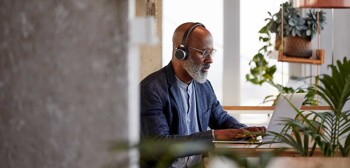 Person wearing headphones sitting at a desk typing on a laptop surrounded by plants Person wearing headphones sitting at a desk typing on a laptop surrounded by plants