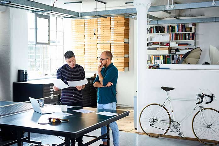 Two people standing in front of a ping pong table covered in paper, one person is holding two sheets of paper which both are looking at pensively  Two people standing in front of a ping pong table covered in paper, one person is holding two sheets of paper which both are looking at pensively