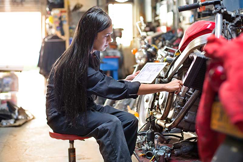 A person sitting on a stool wearing overalls holding a tablet and touching a motorbike in a garage
