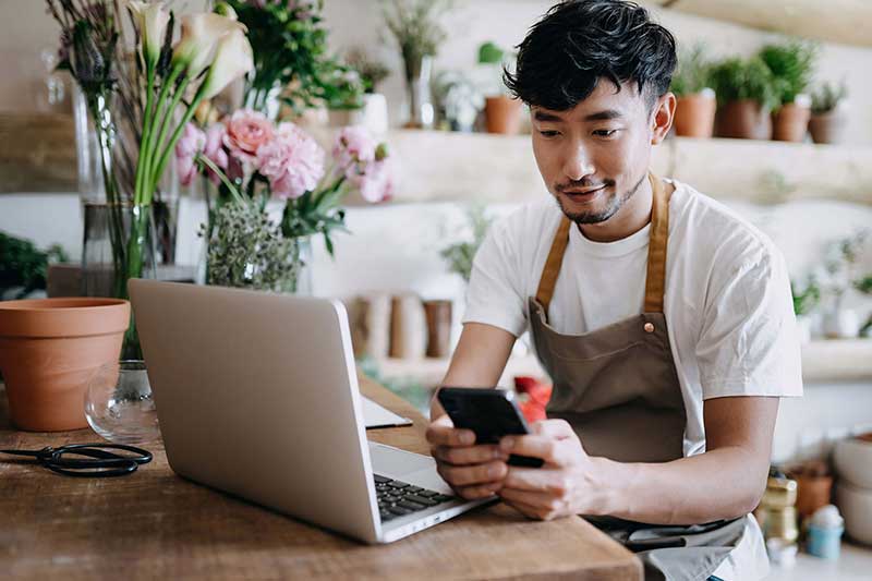 Person wearing an apron sitting in front of a laptop holding their phone in a flower shop