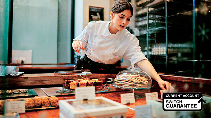 Female baker behind the counter picking up an item to give to a customer Female baker behind the counter picking up an item to give to a customer